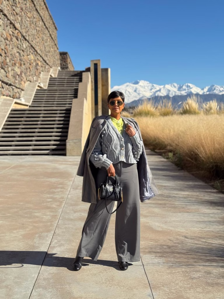 Woman standing outdoors in front of stone stairs and snowy mountains, wearing a gray coat, knit sweater, wide-legged gray pants, and sunglasses, holding a black handbag.